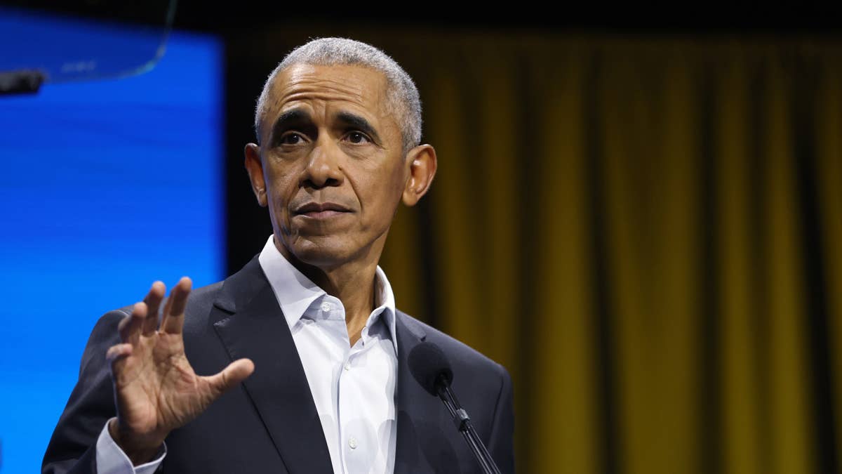 Barack Obama in a dark suit and white shirt speaks at a podium with a blue screen behind him.