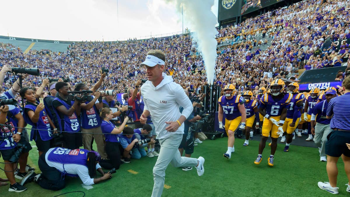 LSU coach Brian Kelly runs onto the field in a white LSU hoodie, with players in purple and gold uniforms behind him.