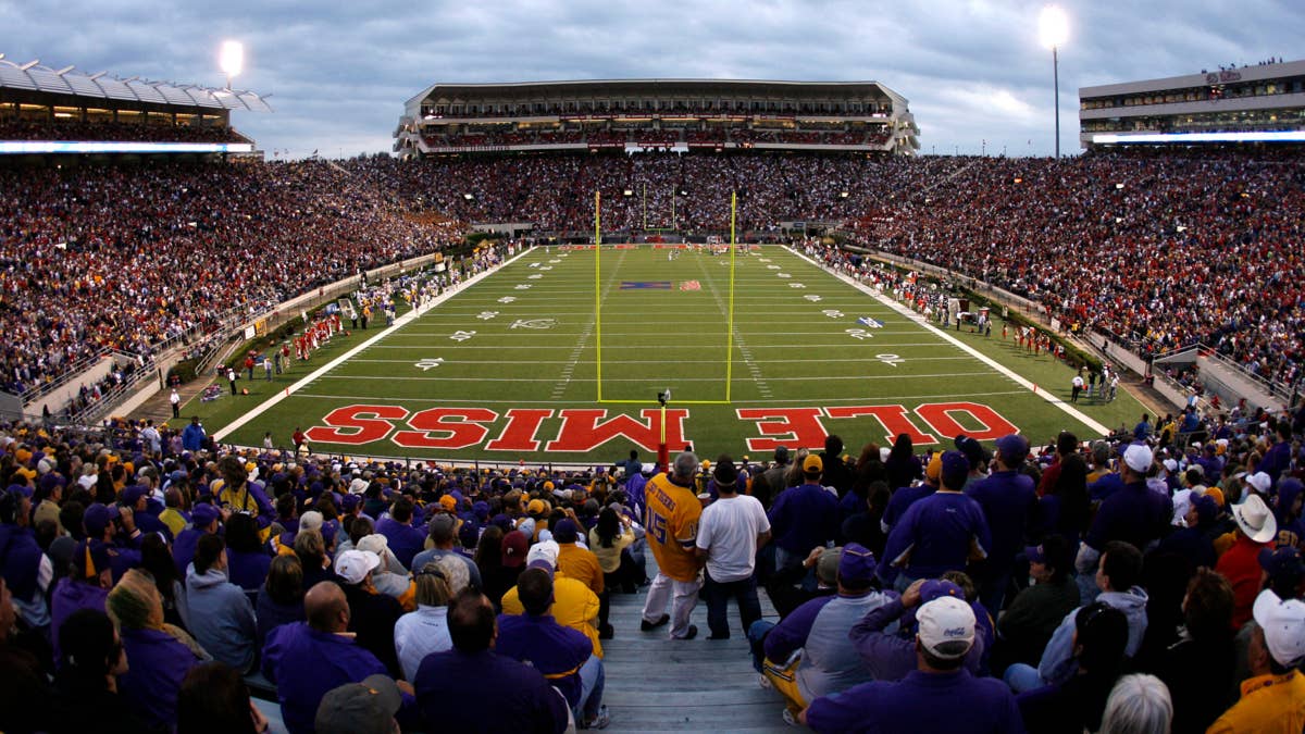 A panoramic view of a football stadium with "OLE MISS" in large red letters on the field, and a crowd of spectators in the stands.