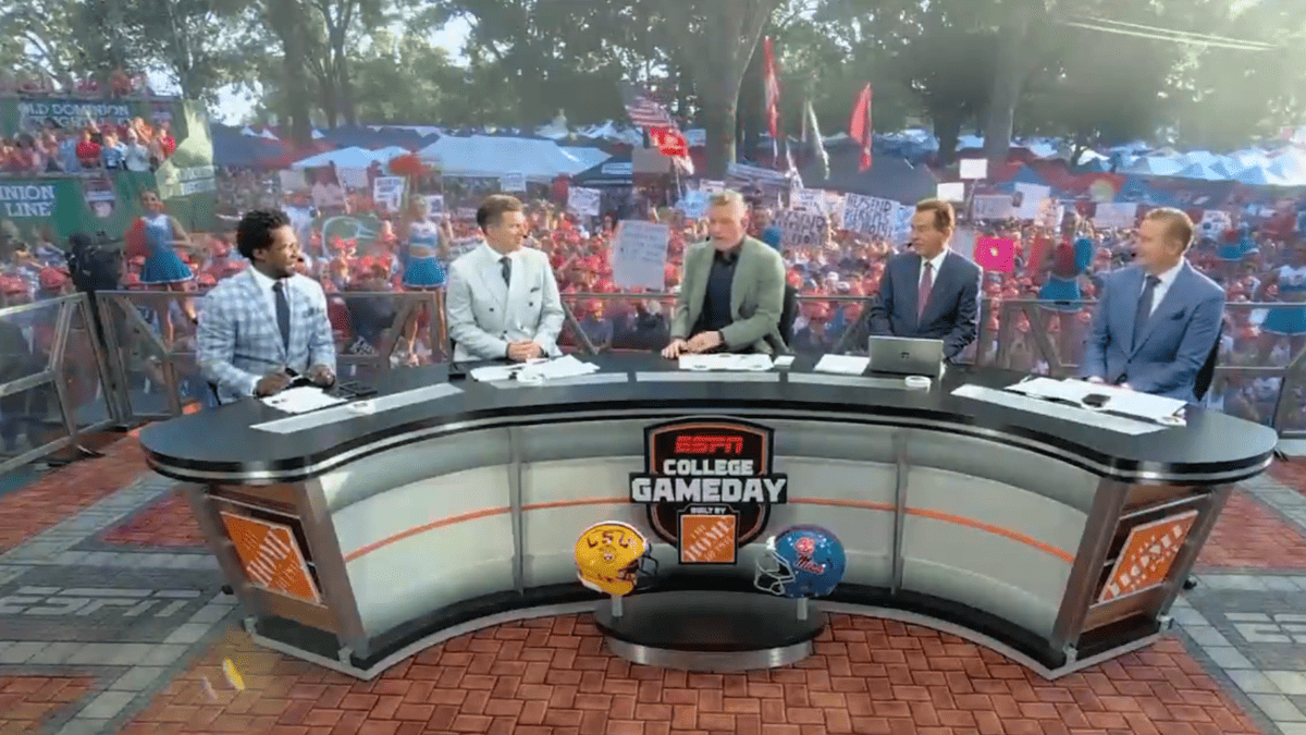 ESPN College GameDay hosts sit at a desk with Ole Miss and LSU helmets in front of a crowd.