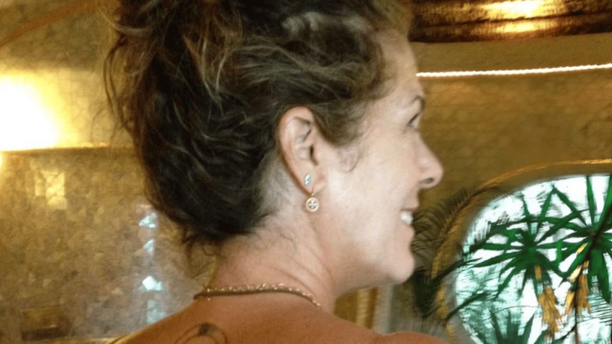 Rita Wilson smiles, wearing a backless swimsuit and earrings, with a stained-glass window behind her.