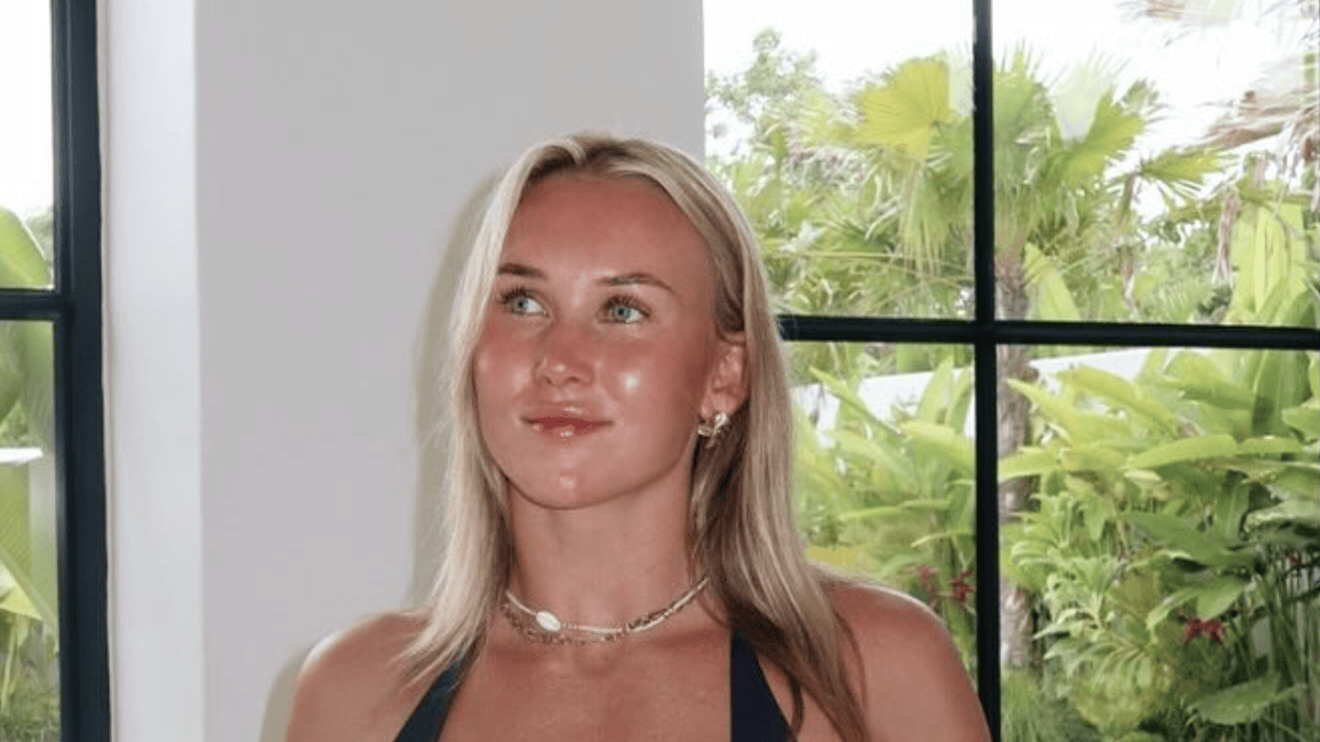 Jamie Chapman in a dark bikini with a shell necklace, looking away from the camera with lush greenery behind her.