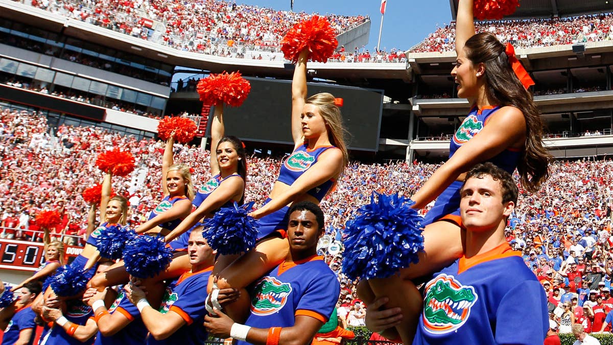 Cheerleaders in blue and orange uniforms with alligator logos hold red and blue pom-poms in a stadium with a large crowd.