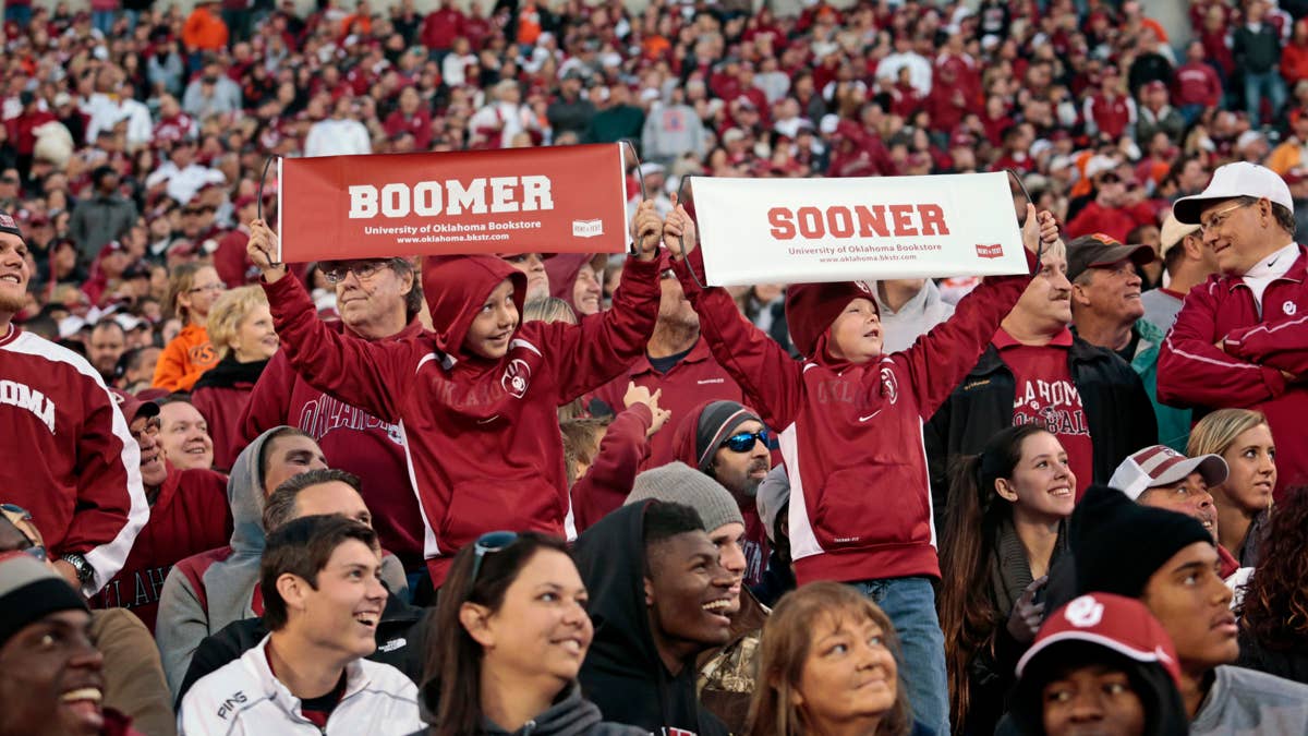 Two children in maroon Oklahoma hoodies hold up signs that say "BOOMER" and "SOONER" in front of a crowd.