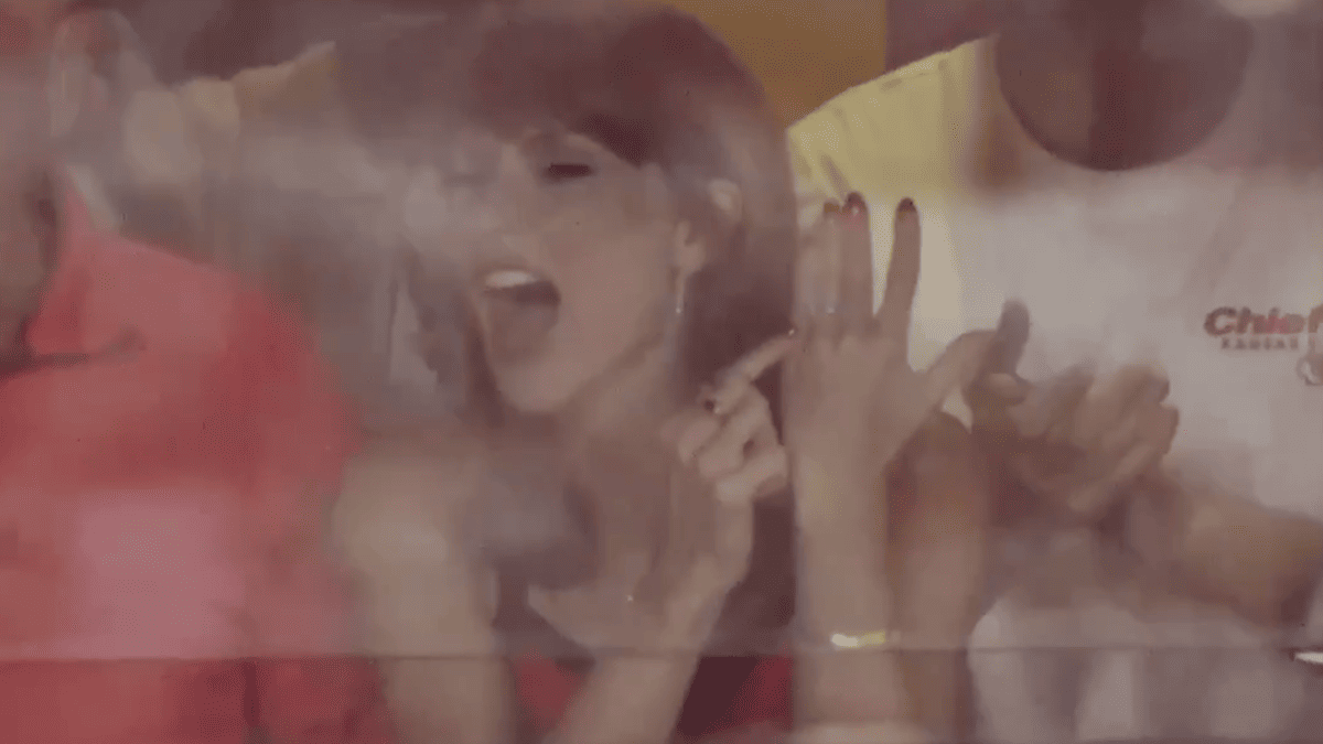 Taylor Swift points to her ring, wearing a Chiefs shirt, during a game.