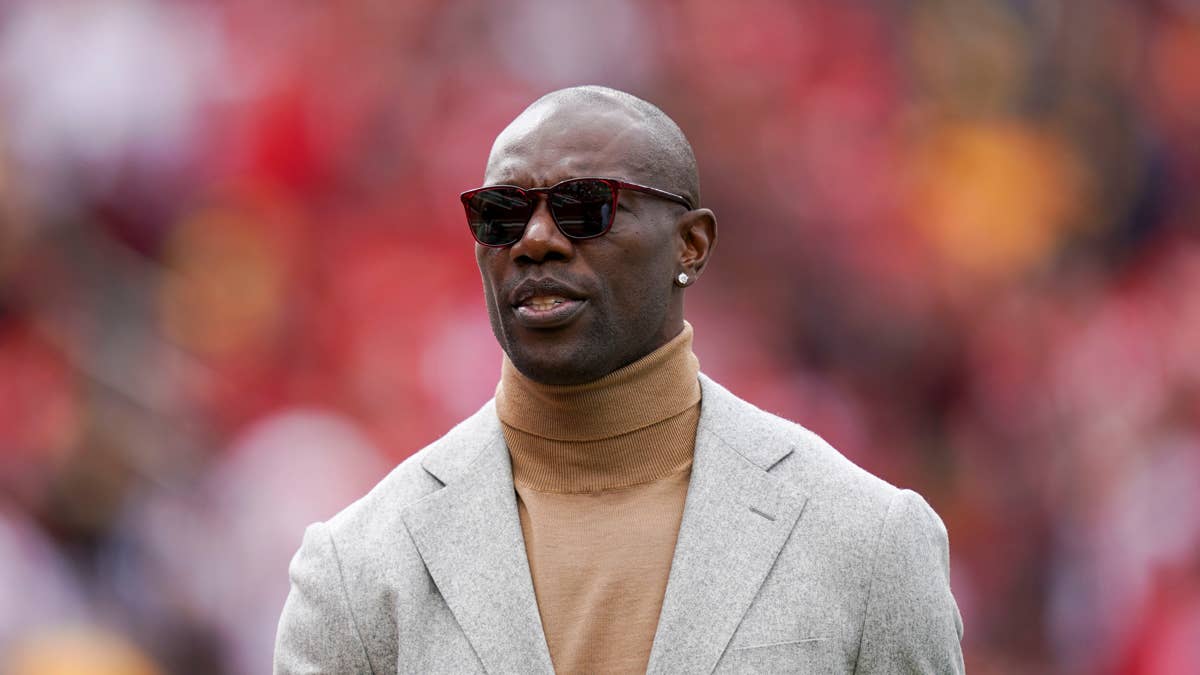 Terrell Owens wears sunglasses, a tan turtleneck, and a gray blazer with a blurred red crowd in the background.