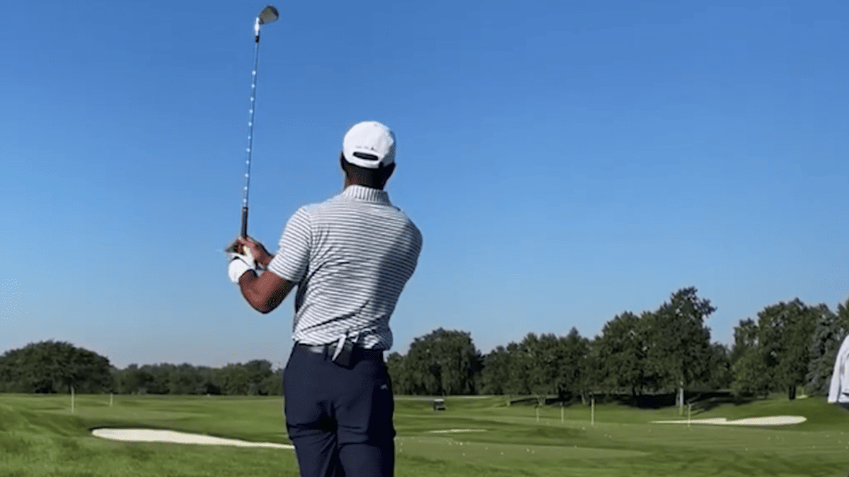 Tiger Woods swings a golf club on a course, wearing a striped shirt and white hat.