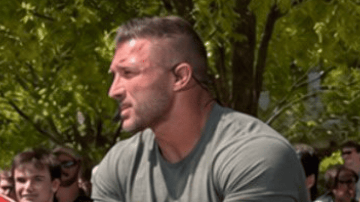 Tim Tebow in a grey t-shirt with a crowd of fans in the background outdoors.