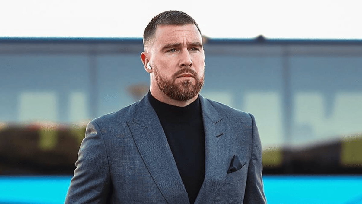 Travis Kelce in a grey suit and black turtleneck, wearing earbuds, before a Chiefs game.