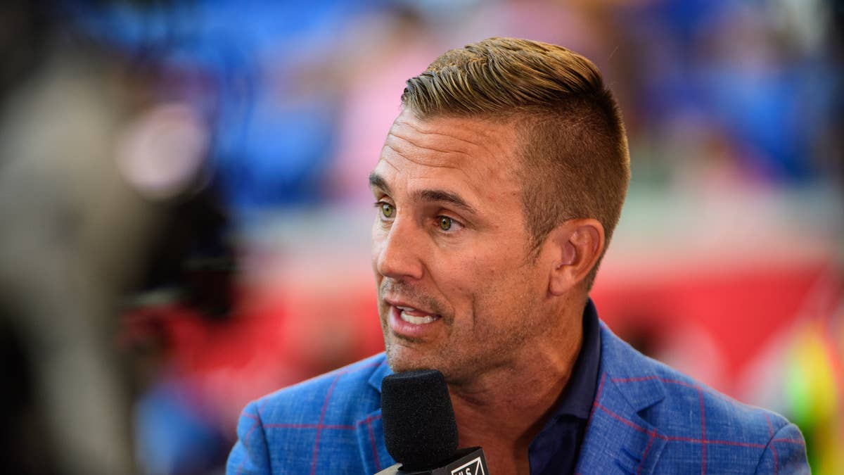 Taylor Twellman speaks into a microphone wearing a blue plaid jacket.