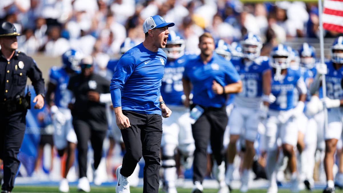 Will Stein wearing a blue shirt and white hat yells as he runs with Kentucky players in blue jerseys.